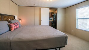 A bedroom with a bed, pillows, a ceiling light, a door leading to a hallway, and a window with blinds. - Red Sands Habitation (Moab)