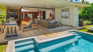 Un espacio exterior moderno con piscina, terraza de madera y cocina con parrilla integrada.