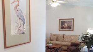 Ein Wohnzimmer mit einer Couch, einem Couchtisch, einem Korbstuhl als Beistelltisch mit Dekoration, einem gerahmten Gemälde und einem Deckenventilator.