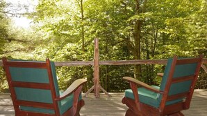 Zwei Adirondack-Stühle mit grünen Kissen auf einer Holzterrasse, umgeben von Bäumen.