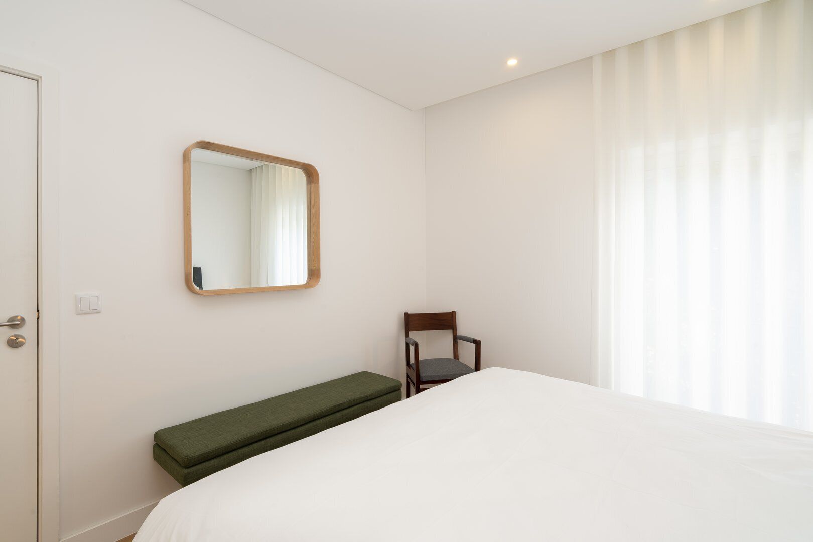 A hotel room with a bed, a bench, a chair, a mirror, and a door.