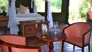 A four-poster bed with a canopy, a wooden chest of drawers, two wine glasses, and two upholstered chairs in a room with wooden flooring and large windows.