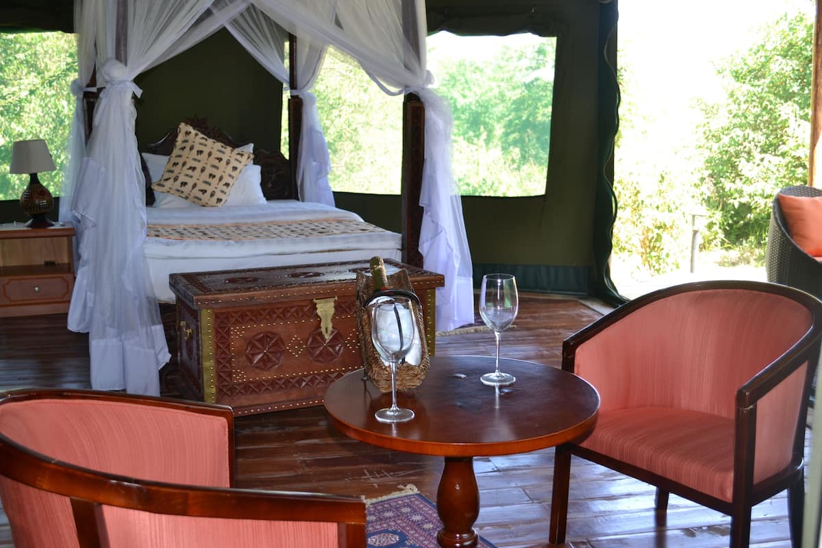 A four-poster bed with a canopy, a wooden chest of drawers, two wine glasses, and two upholstered chairs in a room with wooden flooring and large windows.