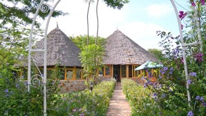 A traditional thatched-roof building with a pathway leading to the entrance, surrounded by lush greenery and flowering plants.