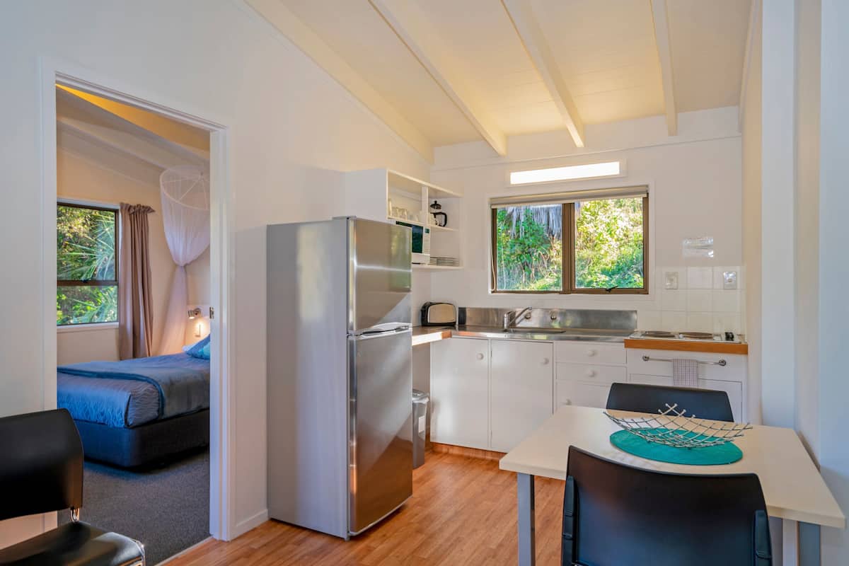 A compact kitchen with a refrigerator, microwave, and sink. There is a small dining table with chairs. A bed is visible in the adjacent room.