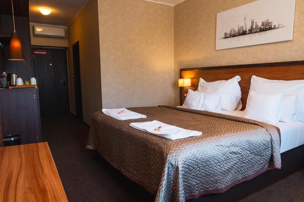 Un cuarto de hotel con una cama, una mesa de madera, una obra de arte con un paisaje urbano en la pared y una puerta al fondo.
