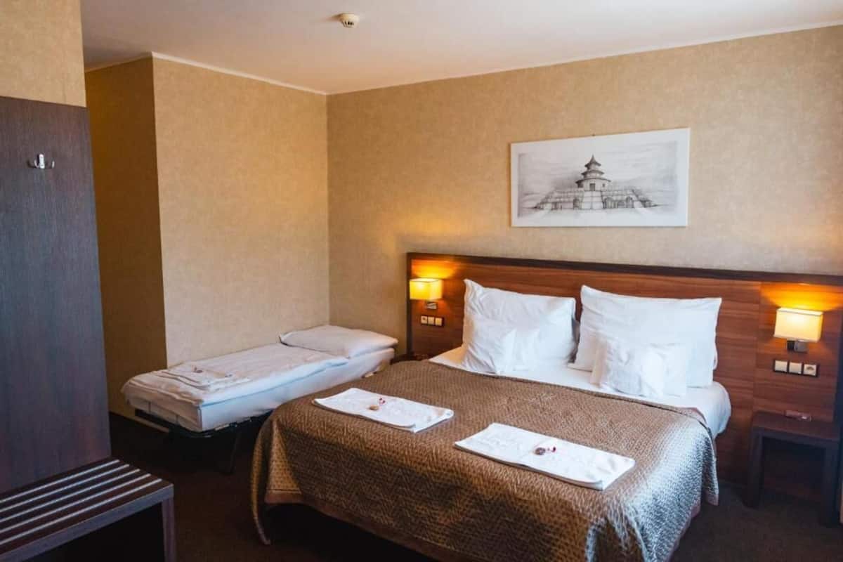 Ein Hotelzimmer mit einem großen Bett, einem kleineren Bett und einem Schreibtisch. An der Wand hängt ein Bild und auf dem Schreibtisch steht eine Lampe.