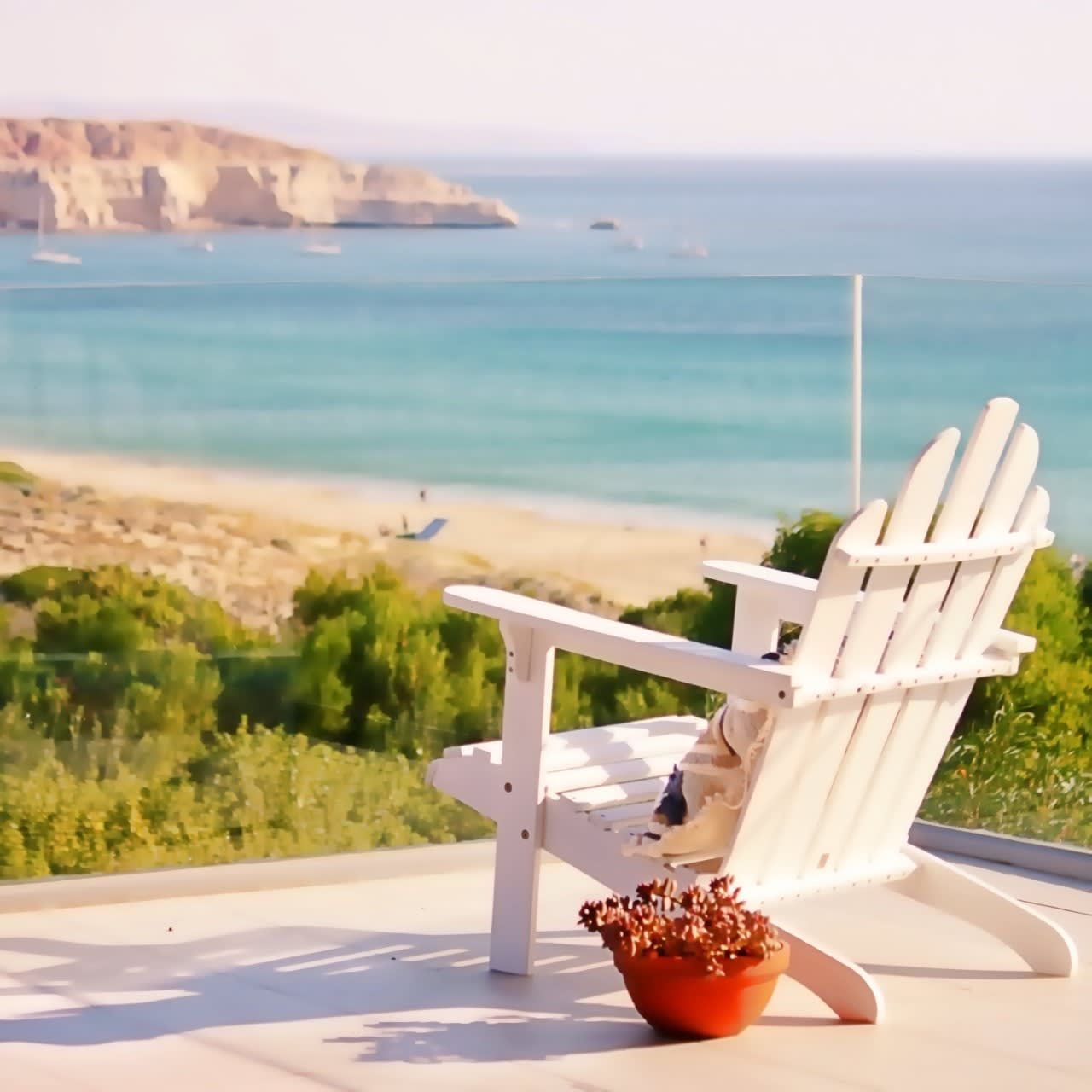 Une chaise Adirondack blanche, ornée d’un pot de fleurs, est placée sur un balcon donnant sur une plage aux eaux d’un bleu limpide et sur des falaises lointaines.