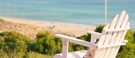 A white Adirondack chair with a potted plant on a balcony overlooking a beach with clear blue waters and distant cliffs.