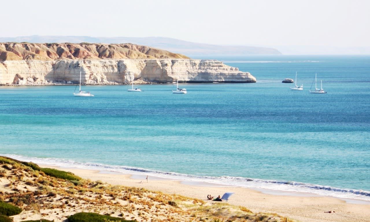 Escena costera con una playa arenosa, acantilados y varios veleros en el agua.