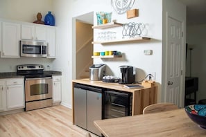 Uma cozinha compacta com micro-ondas, forno e máquina de lavar louça, uma mesa de madeira e um balcão pequeno com cafeteira e chávenas.