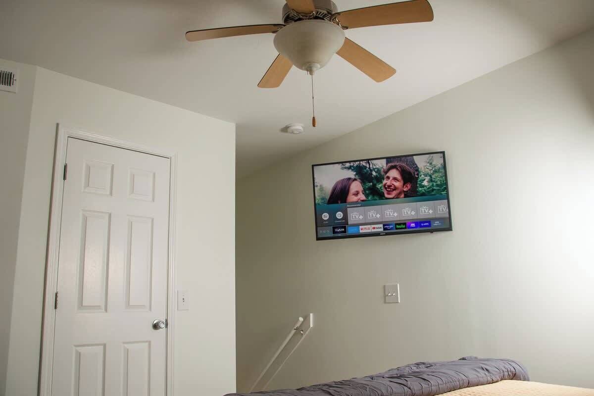 A bedroom with a bed, a ceiling fan, a TV mounted on the wall, and a door.
