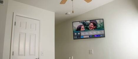 A bedroom with a bed, a ceiling fan, a TV mounted on the wall, and a door.