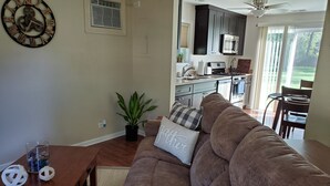 A living room with a brown sofa, a wooden coffee table, and a potted plant. A kitchen is visible in the background with dark cabinets and a microwave. - Cozy Cabin In Roselle (Roselle)