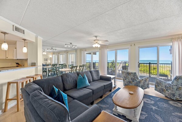 A spacious living area with a gray sofa, a wooden coffee table, and a view of the ocean through large windows. - Destiny: Oceanfront, Beach Access, Shared Pool (Nags Head)