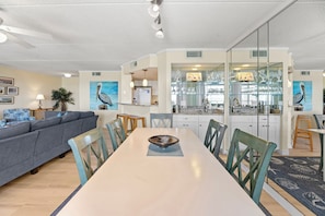 A modern living room with a sofa, dining table, chairs, and a kitchen area with a bar. - Destiny: Oceanfront, Beach Access, Shared Pool (Nags Head)