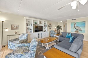 A living room with a grey sofa, patterned armchairs, a coffee table, a TV, and a ceiling fan. - Destiny: Oceanfront, Beach Access, Shared Pool (Nags Head)