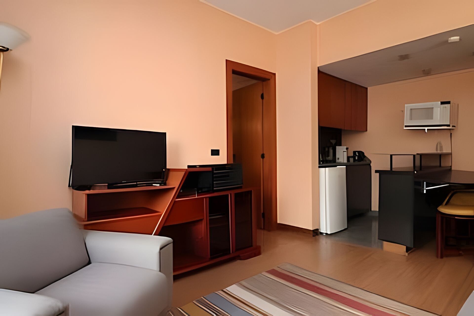 A modern living room with a flat-screen TV, a sofa, a wooden entertainment unit, and a kitchen area with a microwave.