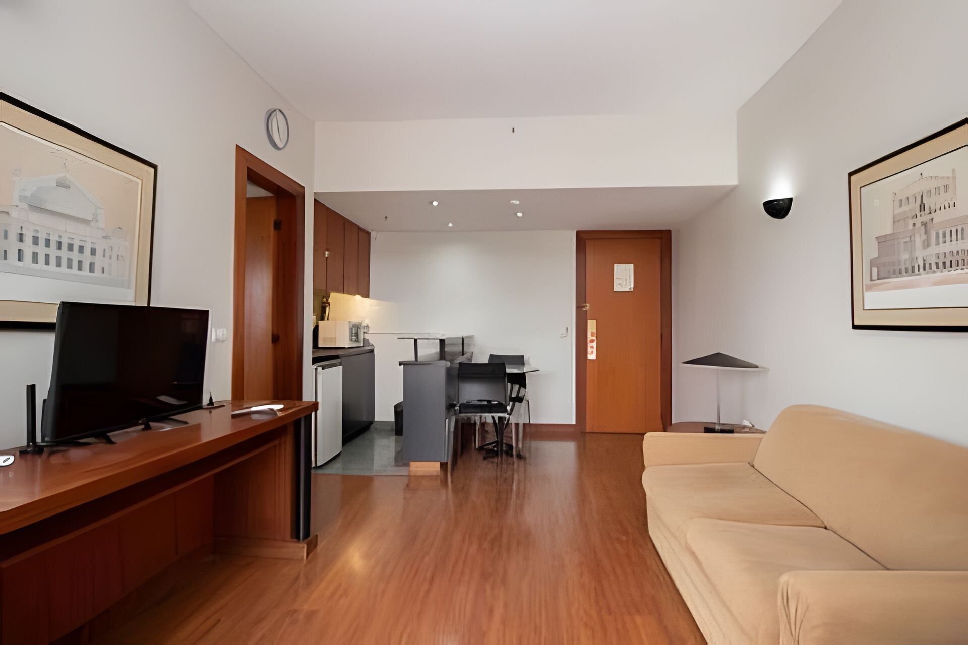 A modern living room with a wooden desk, a television, a sofa, a kitchen area, and framed artwork on the walls.
