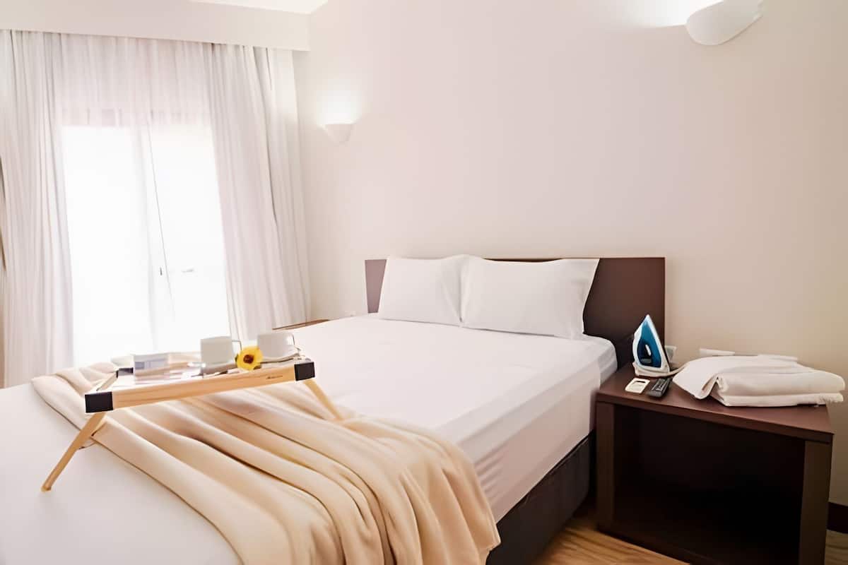Ein Hotelzimmer mit einem Bett, einem Nachttisch mit Bügeleisen, einem Tablett mit einer Tasse und einem Fenster mit Vorhängen.