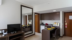 A modern hotel room with a flat-screen TV, a desk with a chair, a large mirror, and a kitchenette. - OZPED Flats Particulares Bonaparte (Brasília)