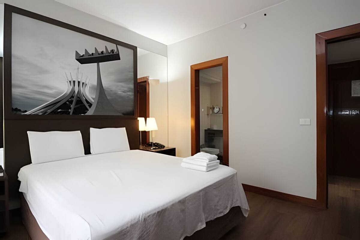 Ein Hotelzimmer mit einem großen Bett, einem Nachttisch mit Lampe, einem Spiegel und einer Tür zu einem anderen Raum.