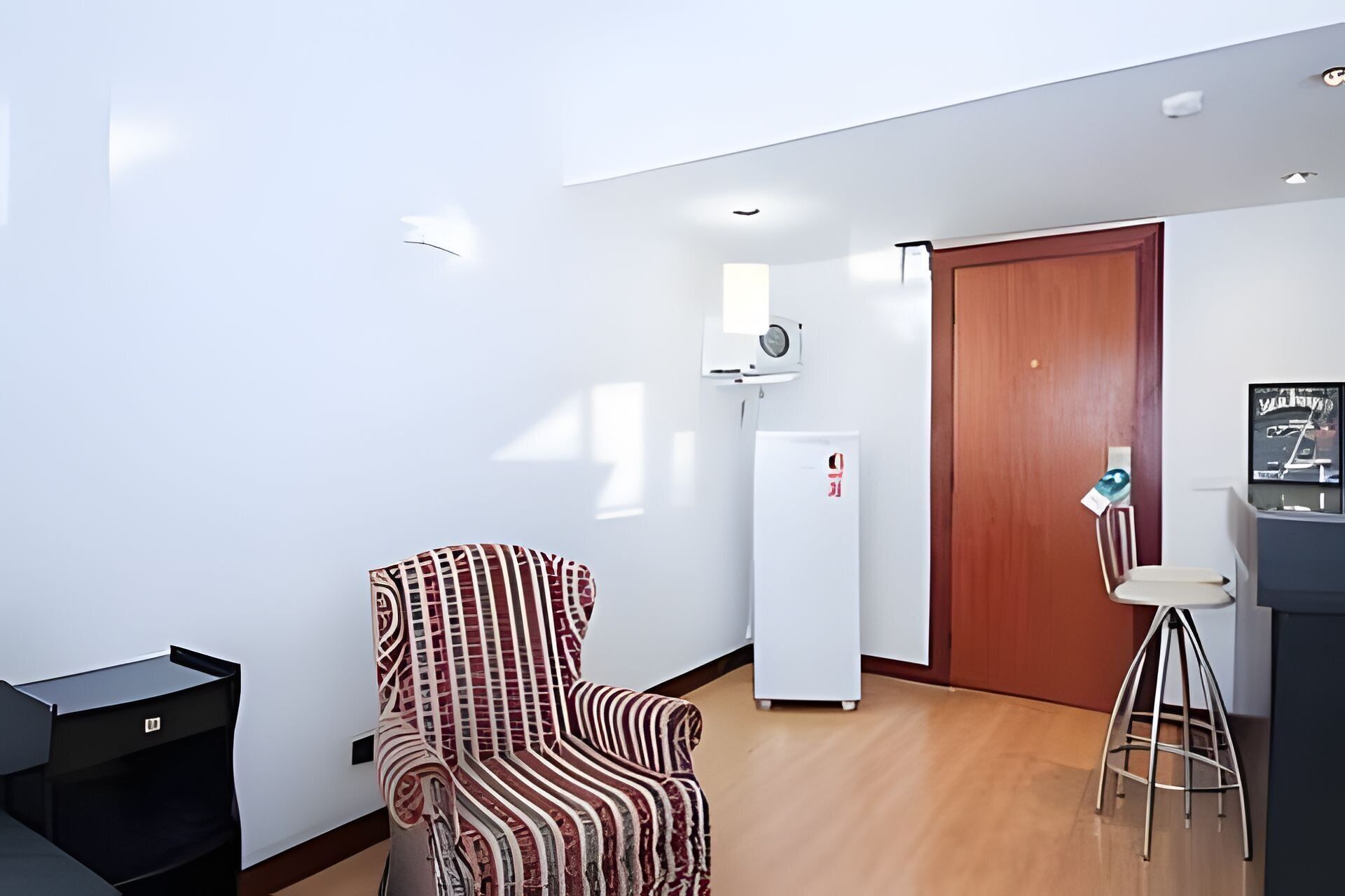 A room with a patterned chair, a white refrigerator, a wooden door, and a small table with a chair.