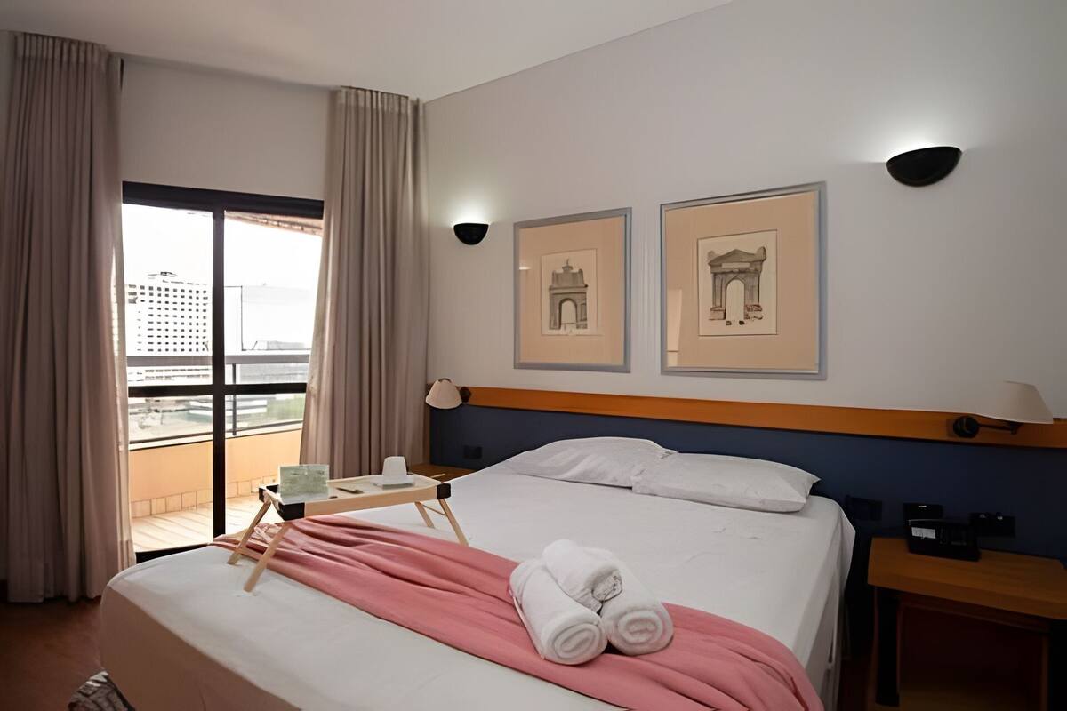 Ein Hotelzimmer mit Bett, Nachttisch, Schreibtisch, Stuhl, einem Fenster mit Vorhängen und zwei gerahmten Bildern an der Wand.