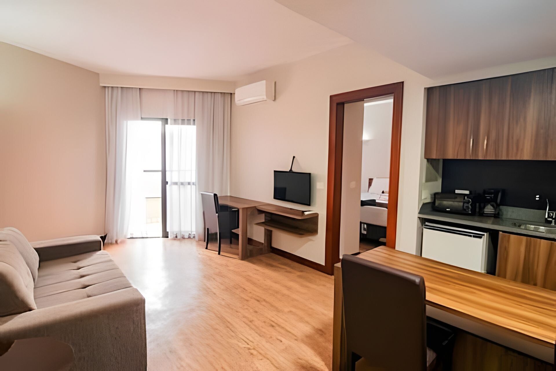 Quarto de hotel moderno com sofá, escrivaninha com computador, televisão, cozinha compacta e porta deslizante que dá para uma varanda.