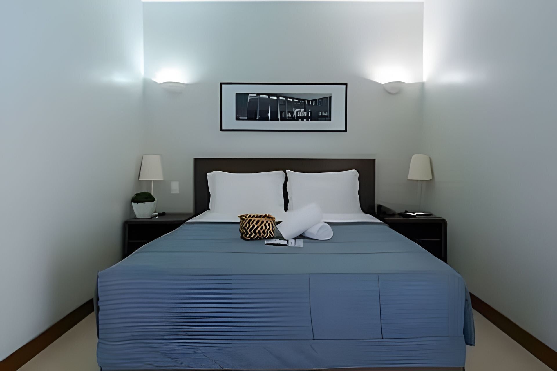 A neatly made bed with a blue bedspread, two white pillows, a decorative pillow, and a small bag on the nightstand.