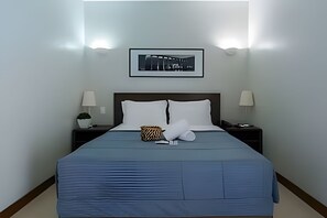 A neatly made bed with a blue bedspread, two white pillows, a decorative pillow, and a small bag on the nightstand. - OZPED Flats Particulares Bonaparte (Brasília)