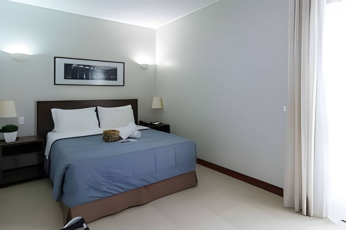 Ein Hotelzimmer mit einem Bett, zwei Nachttischen, einem Fenster mit Vorhängen und einem gerahmten Bild an der Wand.
