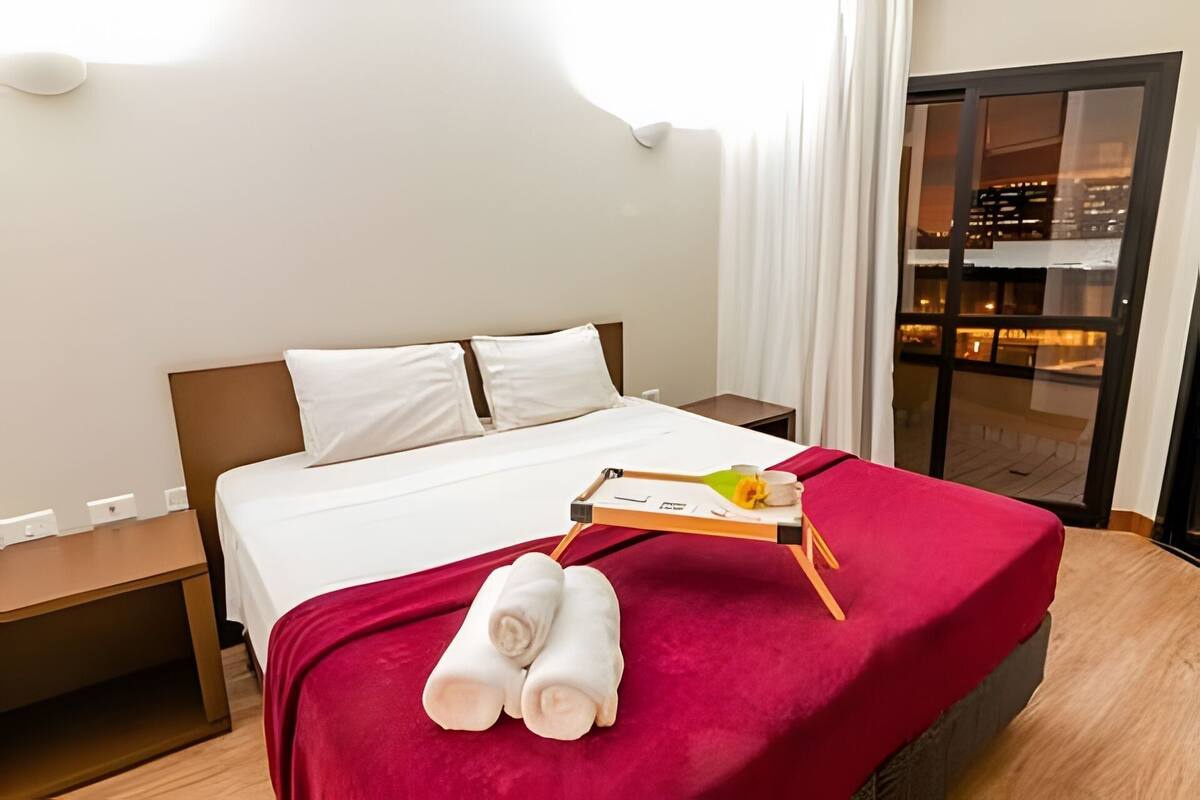 Ein Hotelzimmer mit einem Bett, einem Nachttisch, einer Ablage mit Handtüchern und einem Buch sowie Blick auf die Stadt durch eine Glastür.