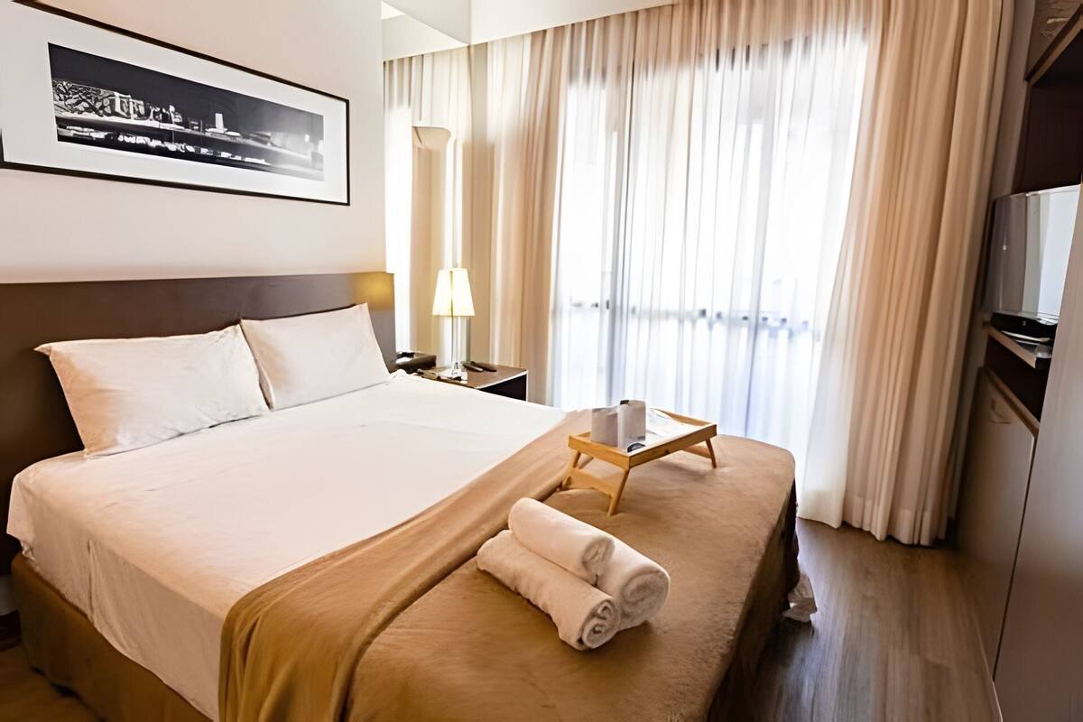 Ein Hotelzimmer mit einem Bett, einem Nachttisch, einer Lampe, einem Fenster mit Vorhängen und einem gerahmten Bild an der Wand.