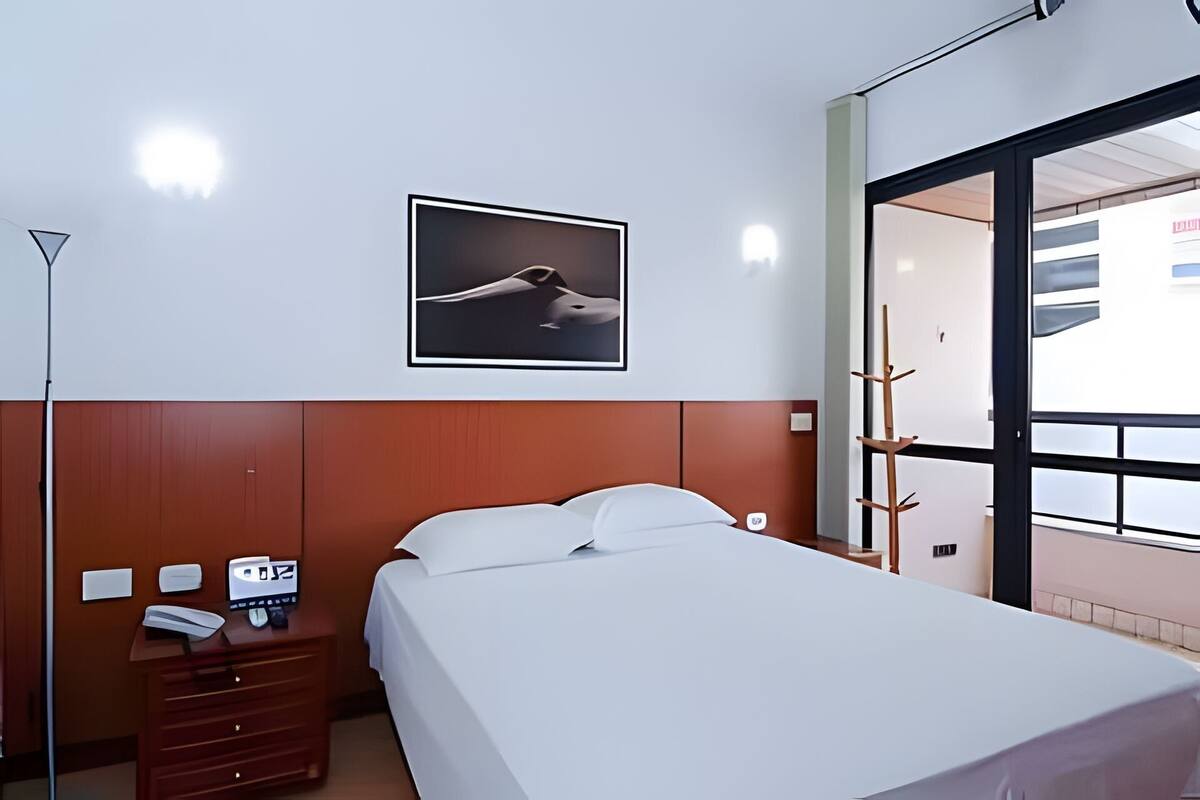 Ein Hotelzimmer mit Bett, Schreibtisch mit Telefon, Kleiderständer und einem gerahmten Bild an der Wand.
