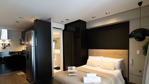 Una habitación de hotel moderna con una cama grande, una mesita de noche pequeña, un escritorio integrado y una kitchenette.