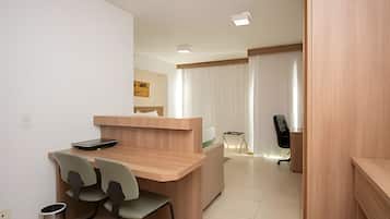 A modern hotel room with a wooden reception desk, two chairs, a small trash bin, and a black office chair.