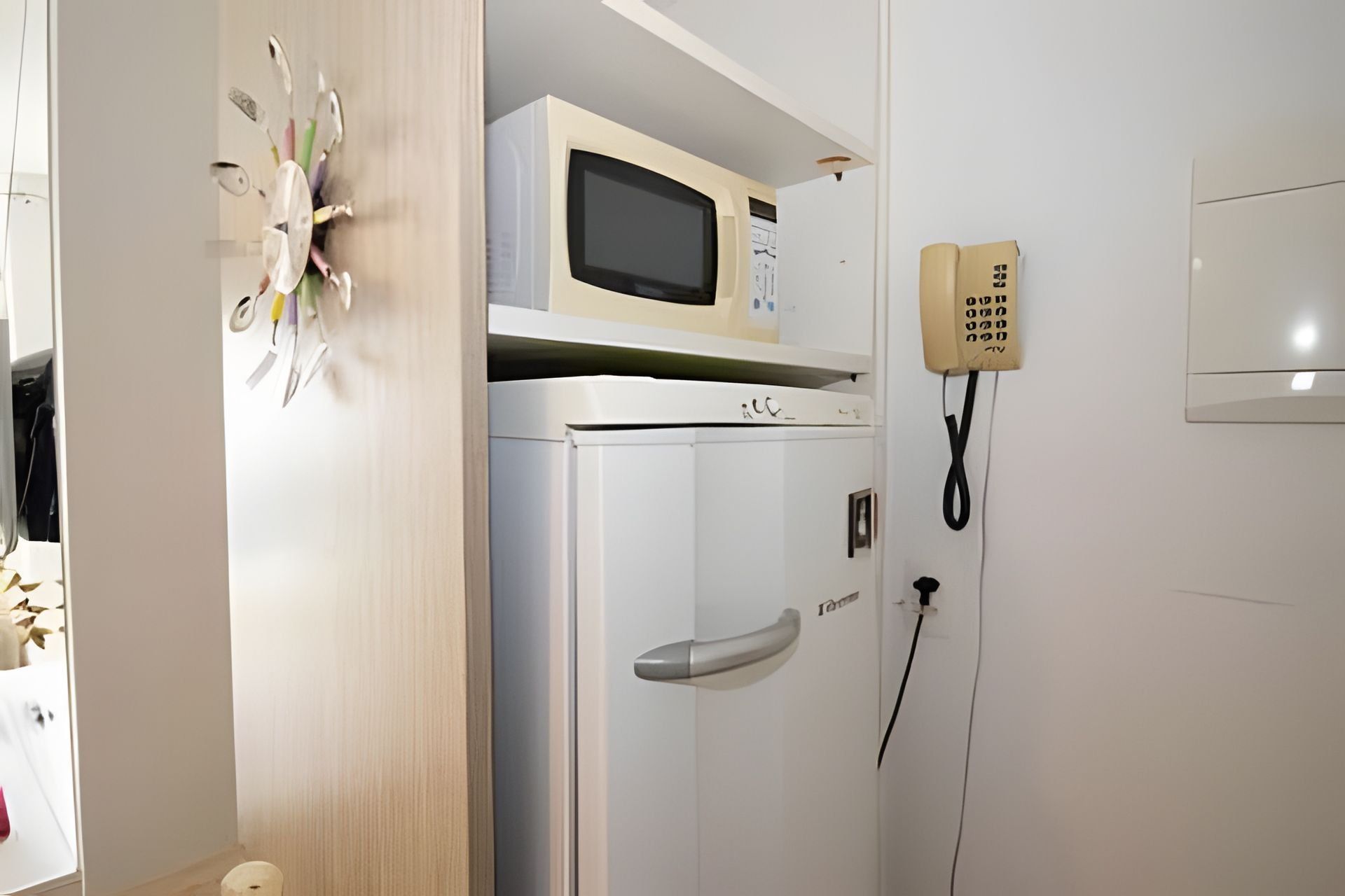 Een kamer met een magnetron bovenop een witte koelkast, een telefoon en een deur met een handvat.