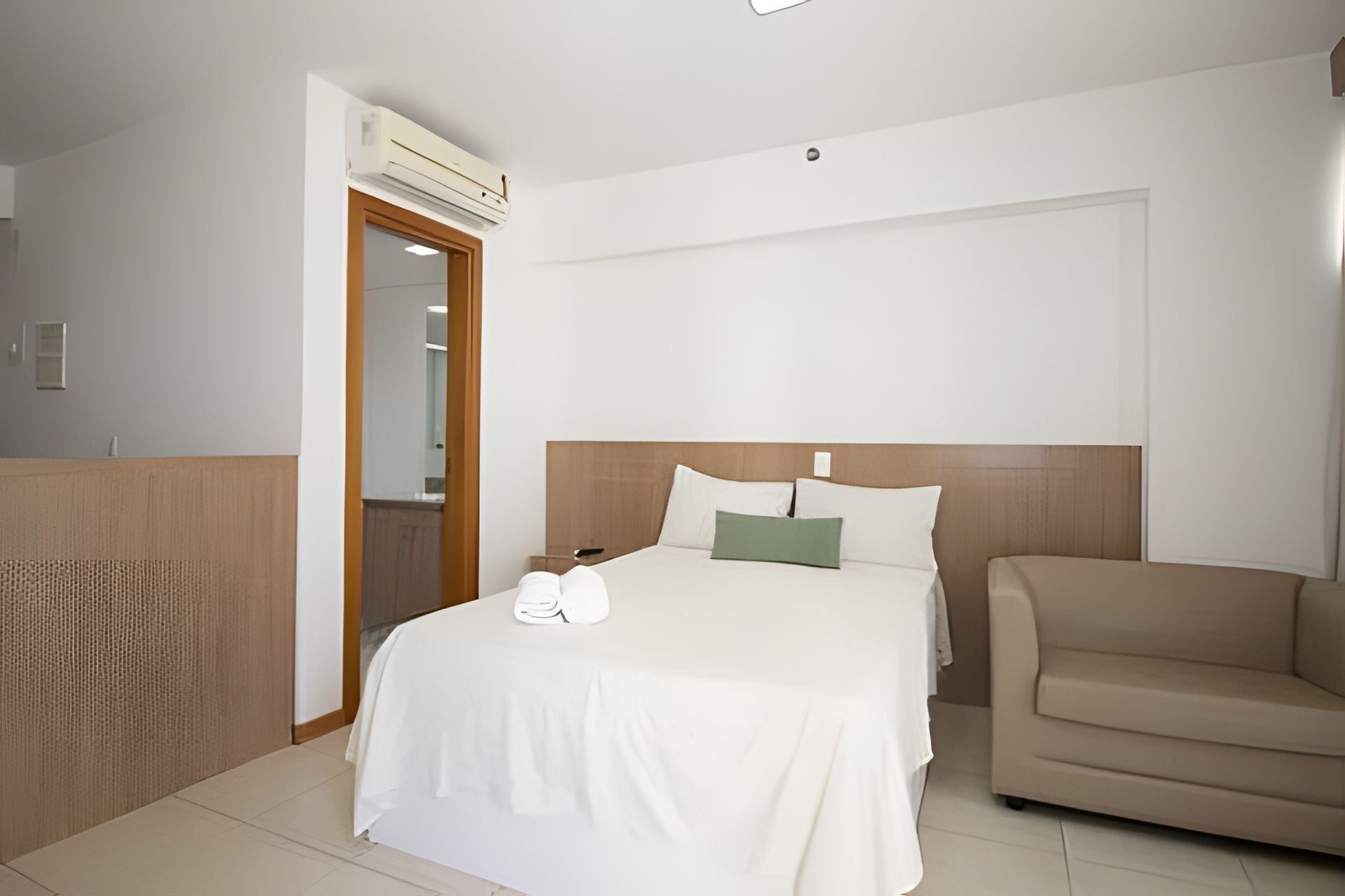 A hotel room with a bed, a chair, a wooden headboard, a door, and an air conditioning unit.