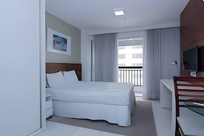 A hotel room with a bed, a desk, a chair, a TV, a painting on the wall, and a balcony with curtains.