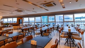Un vaste espace dédié à la salle à manger, avec de nombreuses tables et chaises, de grandes fenêtres offrant une vue sur le paysage urbain et un design moderne au niveau du plafond.