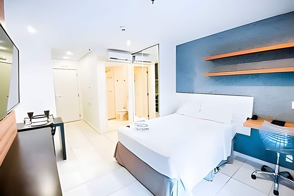 A modern hotel room with a bed, a desk, a mirror, a wardrobe, and a bathroom visible through an open door.