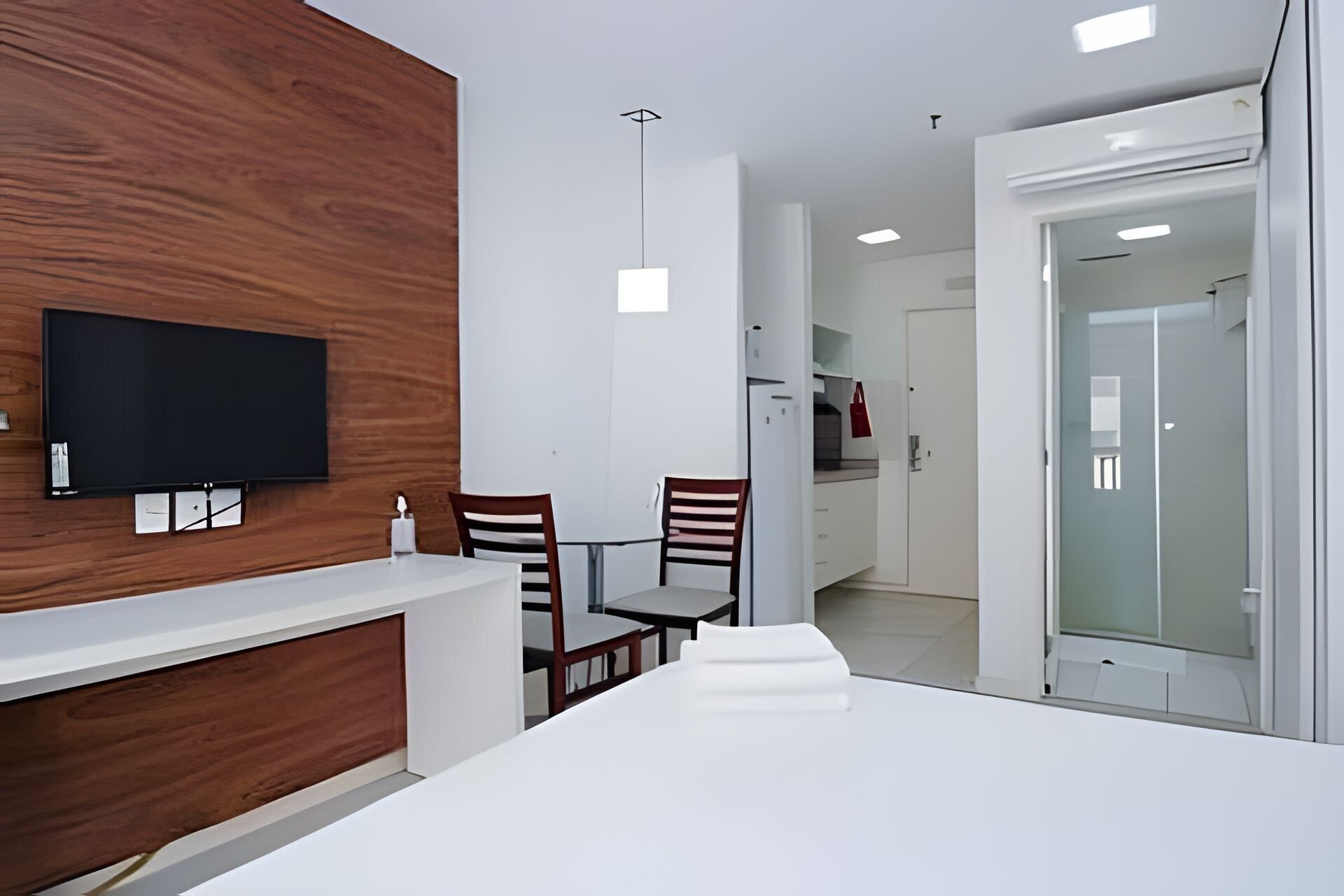 Una habitación moderna con un panel de madera decorativo, un televisor de pantalla plana, una mesa de comedor con sillas y una cocina visible a través de una puerta abierta.