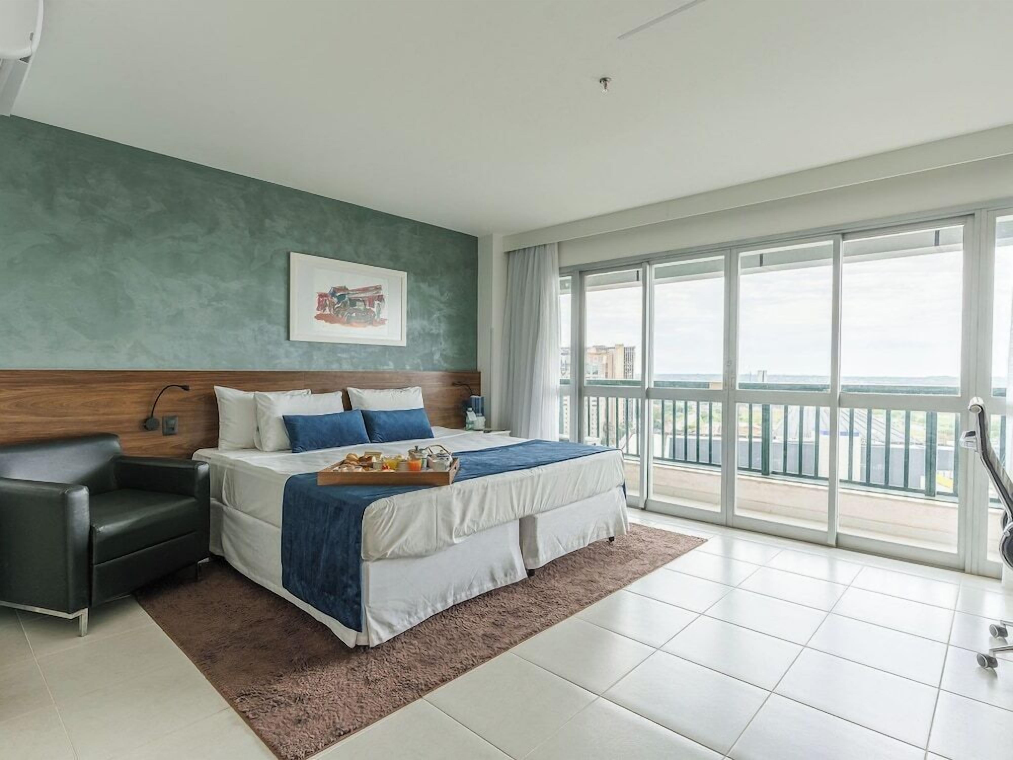 A modern hotel room with a large bed, a bedside table, a chair, and a balcony with a city view.