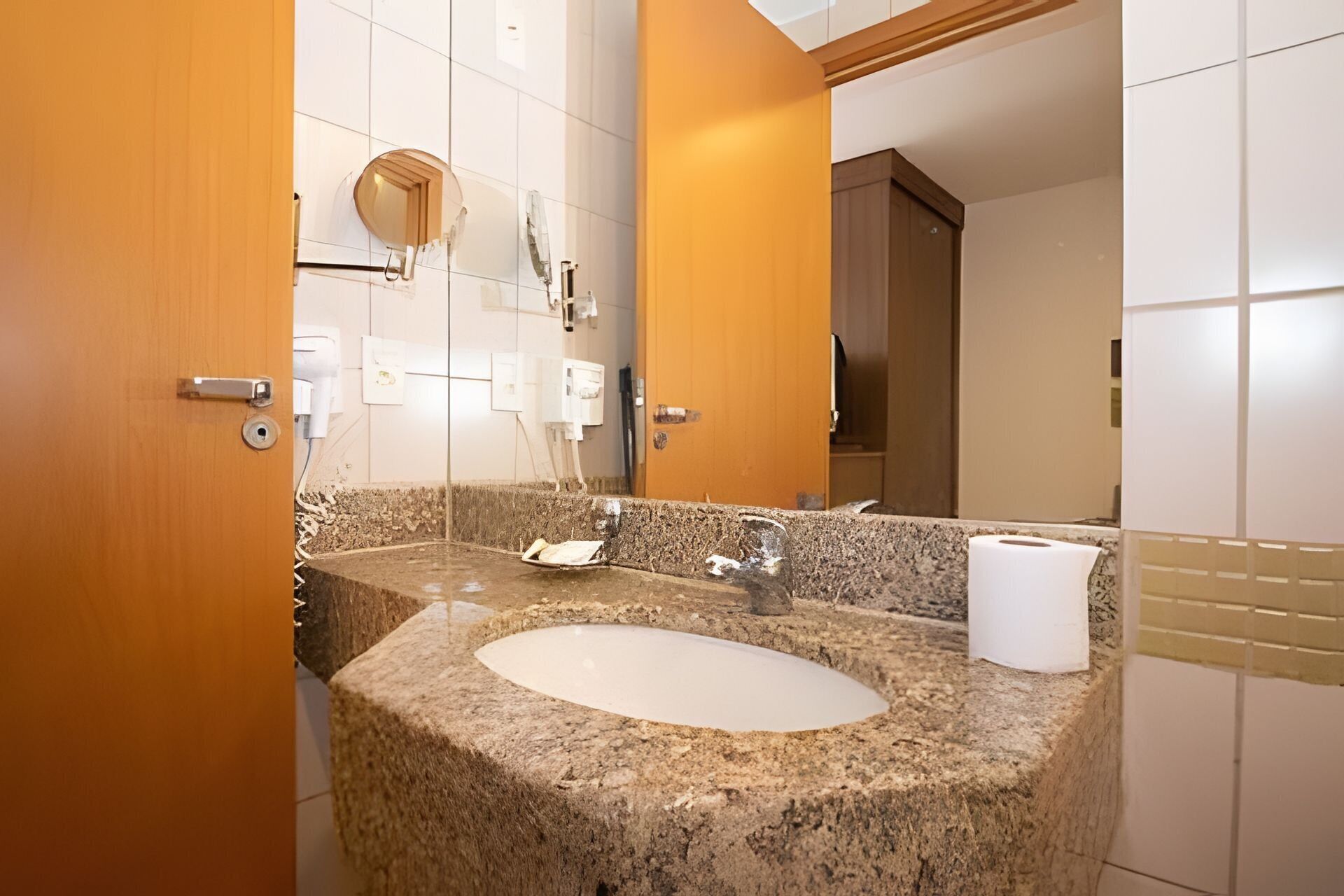 A bathroom with a granite sink, a mirror, a toilet paper dispenser, and a wooden door.