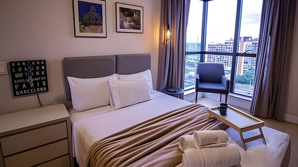 Une chambre d’hôtel avec un grand lit, un bureau, une chaise et une vue sur la ville grâce à de grandes fenêtres.