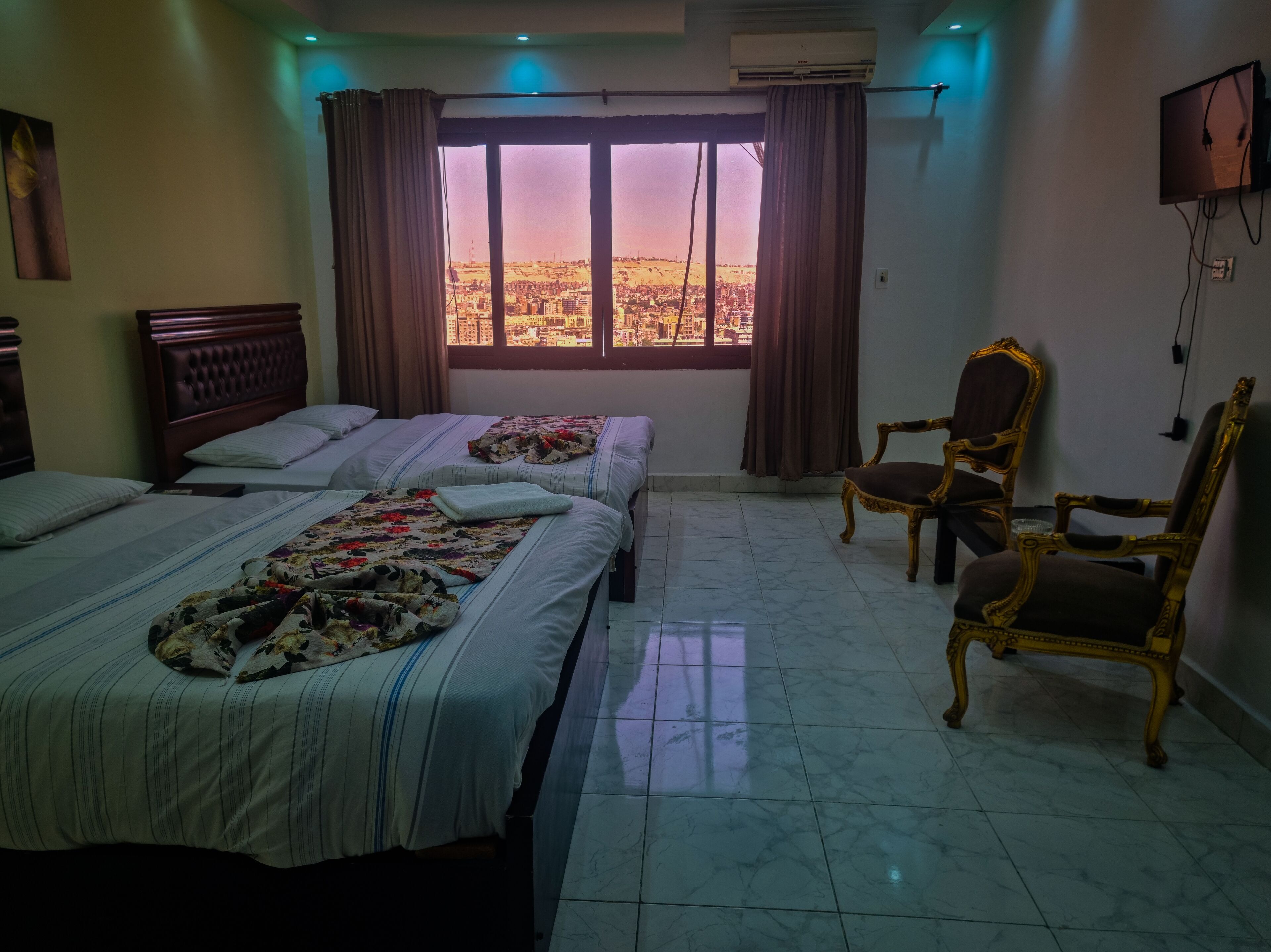 Habitación de hotel con dos camas, un ventanal con cortinas, un televisor montado en la pared y una silla decorativa.