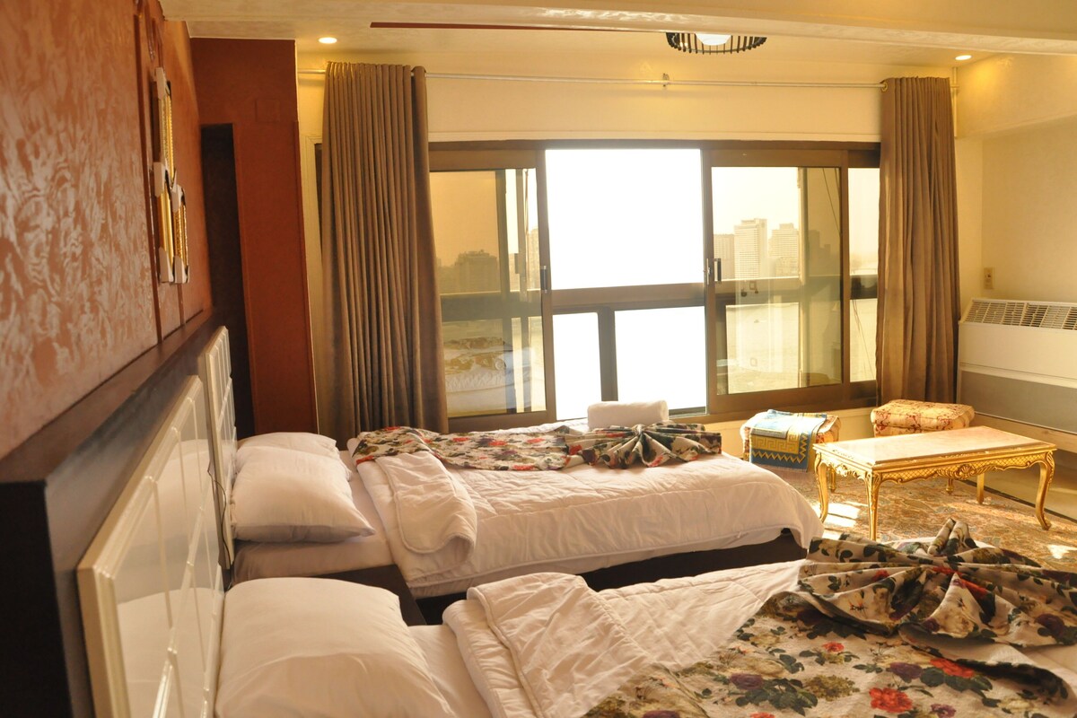 A hotel room with two beds, a small table, and a view of the city through large windows.