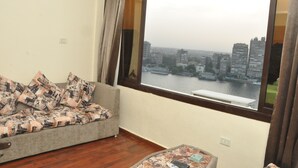 A room with a large window, a grey sofa with patterned cushions, and a small table with a remote control. - Nile sky hotel and suites (Cairo)