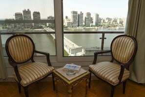 A room with two striped chairs, a small table with a book and ashtray, and a large window overlooking a cityscape and river. - Nile sky hotel and suites (Cairo)